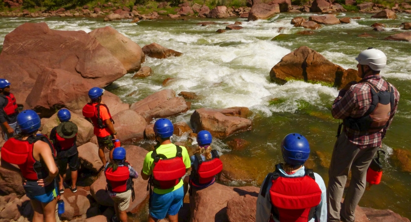 A group of people wearing safety gear stand on the shore of a river, looking out over the water.